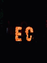Large, illuminated letters spell out 'EC' in a dark environment, with numerous light bulbs embedded in them. A silhouette of a person stands to the right of the letters, partially visible against the bright lighting of the sign.