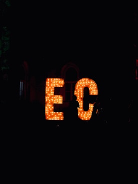 Large, illuminated letters spell out 'EC' in a dark environment, with numerous light bulbs embedded in them. A silhouette of a person stands to the right of the letters, partially visible against the bright lighting of the sign.