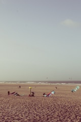 A sandy beach with several kitesurfing sails lying on the ground. In the background, the ocean waves roll towards the shore. Some people are engaged in setting up equipment, while others are walking along the beach. The sky is clear with a few clouds.