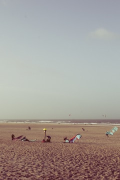 A sandy beach with several kitesurfing sails lying on the ground. In the background, the ocean waves roll towards the shore. Some people are engaged in setting up equipment, while others are walking along the beach. The sky is clear with a few clouds.