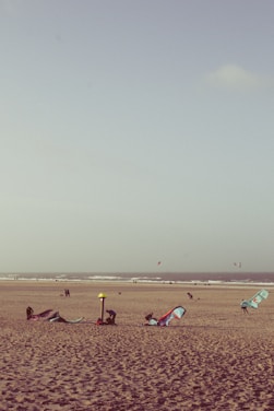 A sandy beach with several kitesurfing sails lying on the ground. In the background, the ocean waves roll towards the shore. Some people are engaged in setting up equipment, while others are walking along the beach. The sky is clear with a few clouds.
