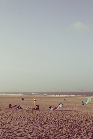 A sandy beach with several kitesurfing sails lying on the ground. In the background, the ocean waves roll towards the shore. Some people are engaged in setting up equipment, while others are walking along the beach. The sky is clear with a few clouds.