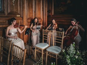 Four musicians in elegant dresses play string instruments in a wooden-paneled room, creating a refined and classical ambiance. The setting includes wooden chairs and floral arrangements, adding to the sophisticated atmosphere.