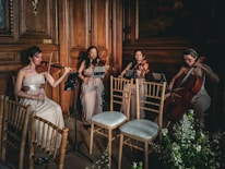 Four musicians in elegant dresses play string instruments in a wooden-paneled room, creating a refined and classical ambiance. The setting includes wooden chairs and floral arrangements, adding to the sophisticated atmosphere.
