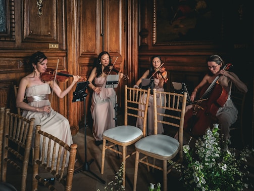 Four musicians in elegant dresses play string instruments in a wooden-paneled room, creating a refined and classical ambiance. The setting includes wooden chairs and floral arrangements, adding to the sophisticated atmosphere.