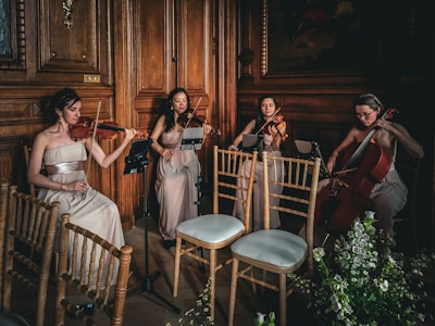 Four musicians in elegant dresses play string instruments in a wooden-paneled room, creating a refined and classical ambiance. The setting includes wooden chairs and floral arrangements, adding to the sophisticated atmosphere.