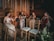 Four musicians in elegant dresses play string instruments in a wooden-paneled room, creating a refined and classical ambiance. The setting includes wooden chairs and floral arrangements, adding to the sophisticated atmosphere.