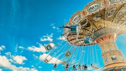 A vibrant amusement park ride featuring a colorful carousel against a bright blue sky scattered with fluffy white clouds. The carousel is intricately decorated, and several blue seats are suspended on long chains, with riders visible, some raising their arms joyously.
