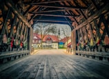 A wooden covered bridge leads to a quaint village with half-timbered houses and a church spire in the background. The bridge interior features decorative garlands and red bows. Trees without foliage are visible outside, suggesting a wintry season.
