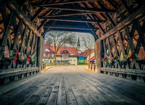 A wooden covered bridge leads to a quaint village with half-timbered houses and a church spire in the background. The bridge interior features decorative garlands and red bows. Trees without foliage are visible outside, suggesting a wintry season.
