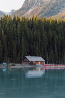 A tranquil lakeside scene featuring a wooden cabin nestled among dense pine trees. The cabin is reflected in the calm turquoise water, with mountains rising in the background. Several red canoes are lined up on a dock beside the cabin.