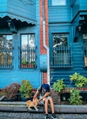 A person wearing a hat is sitting on the curb of a cobblestone street, petting a ginger cat. The background features a blue wooden house with ornate details and a red brick accent, along with pots of greenery and plants.