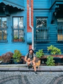 A man is sitting on a cobblestone street in front of a blue wooden house with red brick accents. He is wearing a hat and backpack, smiling with a relaxed posture. Beside him is a ginger cat walking by. The house features ornate iron window grilles and is surrounded by potted plants.