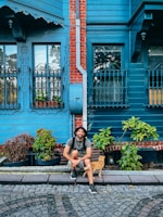 A man is sitting on a cobblestone street in front of a blue wooden house with red brick accents. He is wearing a hat and backpack, smiling with a relaxed posture. Beside him is a ginger cat walking by. The house features ornate iron window grilles and is surrounded by potted plants.