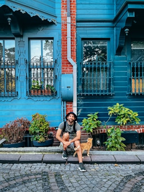 A man is sitting on a cobblestone street in front of a blue wooden house with red brick accents. He is wearing a hat and backpack, smiling with a relaxed posture. Beside him is a ginger cat walking by. The house features ornate iron window grilles and is surrounded by potted plants.