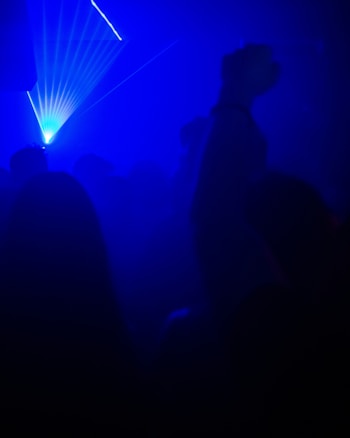 Bright blue laser beams pierce through a dark, smoky environment filled with silhouettes of people. The atmosphere is lively and dynamic, typical of a nightclub or concert scene.
