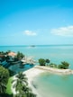 A serene coastal landscape features turquoise waters meeting a clear blue sky. A small island with lush greenery sits amidst calm seas, while palm trees and red-roofed buildings line the shore. The gentle curve of a white sandy beach frames the scene.
