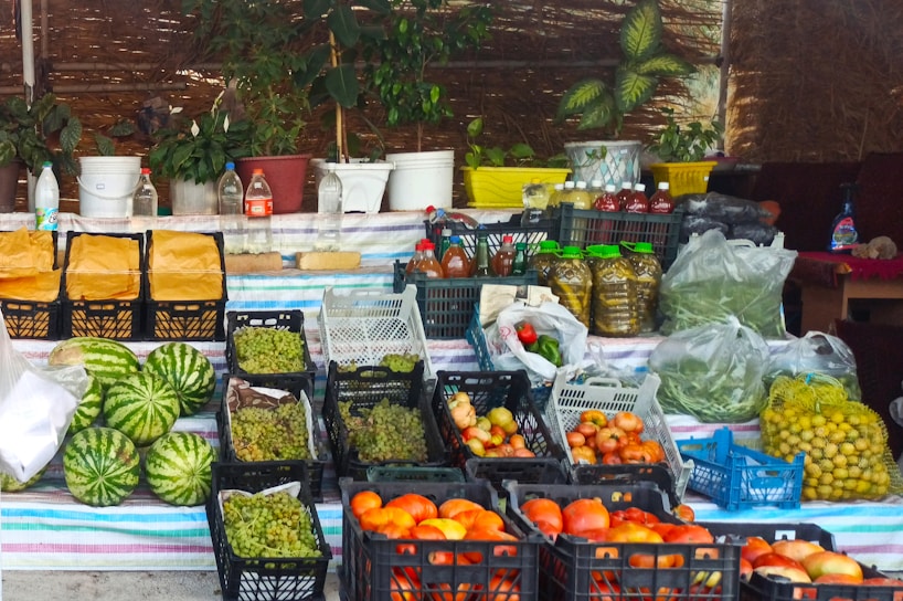 A market stall displaying a variety of fresh produce and other goods. There are several crates of fruits and vegetables, including watermelons, tomatoes, grapes, and apples. The stall also has jars of preserved items, plastic bottles with liquids, and other miscellaneous containers. Plants in pots are placed on the table at the back, and woven screens create a rustic backdrop.