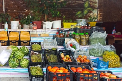 A market stall displaying a variety of fresh produce and other goods. There are several crates of fruits and vegetables, including watermelons, tomatoes, grapes, and apples. The stall also has jars of preserved items, plastic bottles with liquids, and other miscellaneous containers. Plants in pots are placed on the table at the back, and woven screens create a rustic backdrop.