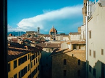 A scenic view of an Italian city with historical architecture, featuring red-tiled rooftops and the prominent dome of a cathedral against a clear blue sky.