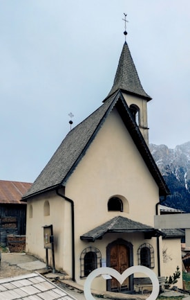 A quaint, small church with beige walls and a steep gray shingle roof, featuring a cross-topped steeple. The building has arched windows and a wooden door. Surrounding the church are rustic elements, including a wooden structure with a rusted roof and mountainous landscape in the background.