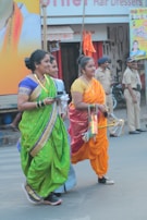 Three women dressed in colorful traditional saris are walking on a street during a daytime event. Two of them are wearing sneakers, and one is holding a set of decorative sticks. In the background, there are police officers and various shop signs.