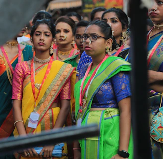 A group of women dressed in colorful traditional attire, including sarees, featuring vibrant colors like yellow, green, and blue. They accessorize with jewelry, such as necklaces and earrings. Some of them wear spectacles and badges around their necks. The background is blurred, suggesting a lively outdoor setting.