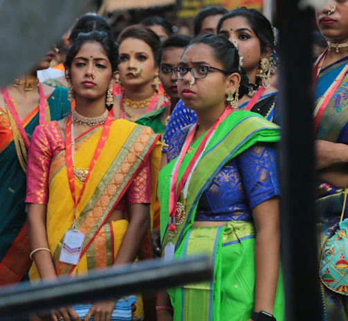 A group of women dressed in colorful traditional attire, including sarees, featuring vibrant colors like yellow, green, and blue. They accessorize with jewelry, such as necklaces and earrings. Some of them wear spectacles and badges around their necks. The background is blurred, suggesting a lively outdoor setting.