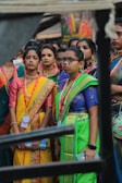 A group of women dressed in colorful traditional attire, including sarees, featuring vibrant colors like yellow, green, and blue. They accessorize with jewelry, such as necklaces and earrings. Some of them wear spectacles and badges around their necks. The background is blurred, suggesting a lively outdoor setting.