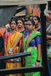 A group of women dressed in colorful traditional attire, including sarees, featuring vibrant colors like yellow, green, and blue. They accessorize with jewelry, such as necklaces and earrings. Some of them wear spectacles and badges around their necks. The background is blurred, suggesting a lively outdoor setting.