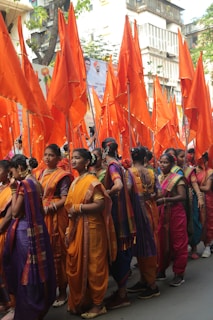 A group of women dressed in colorful traditional outfits participates in a parade. They are holding vibrant orange flags, and are adorned with various pieces of jewelry. The surroundings include banners with images and writing, and the scene appears to be in an urban area.