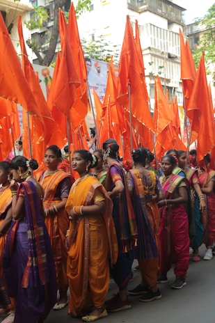 A group of women dressed in colorful traditional outfits participates in a parade. They are holding vibrant orange flags, and are adorned with various pieces of jewelry. The surroundings include banners with images and writing, and the scene appears to be in an urban area.