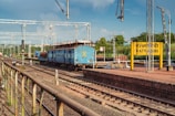 A blue train is stationed on the railway tracks near a platform with a yellow sign reading 'RATNAGIRI' in English and another language. Overhead electric lines are visible, along with metal poles and lights. The background shows greenery and a few buildings under a blue, partly cloudy sky.