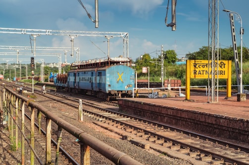 A blue train is stationed on the railway tracks near a platform with a yellow sign reading 'RATNAGIRI' in English and another language. Overhead electric lines are visible, along with metal poles and lights. The background shows greenery and a few buildings under a blue, partly cloudy sky.