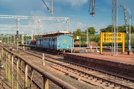 A blue train is stationed on the railway tracks near a platform with a yellow sign reading 'RATNAGIRI' in English and another language. Overhead electric lines are visible, along with metal poles and lights. The background shows greenery and a few buildings under a blue, partly cloudy sky.