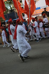 A group of individuals in traditional attire are marching on a street, holding large orange flags. They are dressed in white with red turbans and scarves, appearing to be part of a cultural procession. Crowds line the street, watching the event unfold.