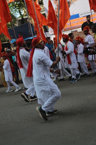 A group of individuals in traditional attire are marching on a street, holding large orange flags. They are dressed in white with red turbans and scarves, appearing to be part of a cultural procession. Crowds line the street, watching the event unfold.