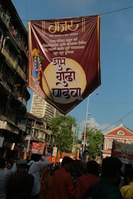 A large banner with Marathi text is hanging above a busy street filled with people. The sky is clear and blue, and the buildings lining the street are a mix of old architecture and newer designs. The crowd below is vibrant, with many people holding orange flags and wearing traditional clothing. The general atmosphere suggests a festival or celebration.
