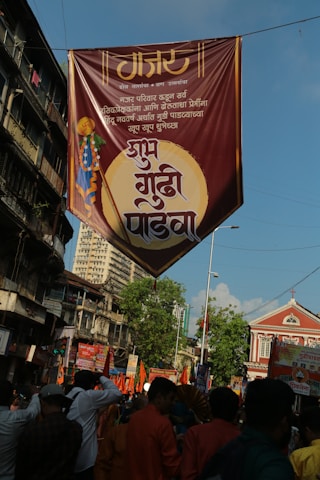 A large banner with Marathi text is hanging above a busy street filled with people. The sky is clear and blue, and the buildings lining the street are a mix of old architecture and newer designs. The crowd below is vibrant, with many people holding orange flags and wearing traditional clothing. The general atmosphere suggests a festival or celebration.