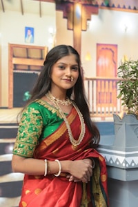 A woman wearing traditional attire featuring a red and green sari with intricate gold embroidery. She accessorizes with gold jewelry, including a necklace and bangles. The background suggests an indoor setting with traditional Indian architectural elements and a potted plant.