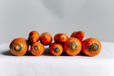 A group of fresh, whole carrots arranged on a white cloth surface with a smooth, gray background. The carrots are bright orange with greenish tops and have a glossy appearance, suggesting they have been washed and are ready for use.