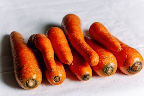 A close-up view of seven fresh, unpeeled carrots lying on a light-colored cloth. The carrots vary in size and shape, exhibiting a vibrant orange color with earthy green tops.