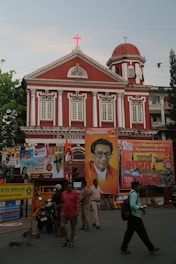 A red building with classical architectural elements stands prominently in the background. It displays a bright red cross atop its facade. In front of the building, several large colorful posters, including a portrait of a man wearing glasses and dressed in traditional attire, are prominently displayed. A group of people, including a person in uniform, walk or stand near the posters. The scene is lively with a sense of activity.