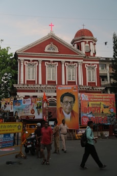 A red building with classical architectural elements stands prominently in the background. It displays a bright red cross atop its facade. In front of the building, several large colorful posters, including a portrait of a man wearing glasses and dressed in traditional attire, are prominently displayed. A group of people, including a person in uniform, walk or stand near the posters. The scene is lively with a sense of activity.