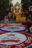 A large, elaborately decorated statue of a deity with a golden face and ornate headdress is placed prominently on a street. The area is adorned with vibrant rangoli designs on the ground featuring intricate geometric patterns and text in a local language. A police barricade from the Mumbai Police station is visible, indicating a controlled area. People are seen walking around, including someone actively working on the rangoli.