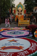 A large, elaborately decorated statue of a deity with a golden face and ornate headdress is placed prominently on a street. The area is adorned with vibrant rangoli designs on the ground featuring intricate geometric patterns and text in a local language. A police barricade from the Mumbai Police station is visible, indicating a controlled area. People are seen walking around, including someone actively working on the rangoli.