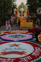 A large, elaborately decorated statue of a deity with a golden face and ornate headdress is placed prominently on a street. The area is adorned with vibrant rangoli designs on the ground featuring intricate geometric patterns and text in a local language. A police barricade from the Mumbai Police station is visible, indicating a controlled area. People are seen walking around, including someone actively working on the rangoli.
