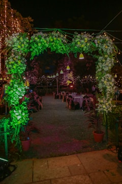 A beautifully decorated outdoor event space at night, featuring a floral archway adorned with lush greenery and white flowers. Behind the archway, tables are set up with chairs, each dressed with dark covers tied with bows. The atmosphere is festive, highlighted by string lights and hanging lamps casting a warm glow.