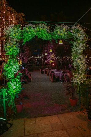 A beautifully decorated outdoor event space at night, featuring a floral archway adorned with lush greenery and white flowers. Behind the archway, tables are set up with chairs, each dressed with dark covers tied with bows. The atmosphere is festive, highlighted by string lights and hanging lamps casting a warm glow.
