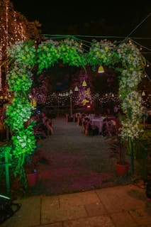 A beautifully decorated outdoor event space at night, featuring a floral archway adorned with lush greenery and white flowers. Behind the archway, tables are set up with chairs, each dressed with dark covers tied with bows. The atmosphere is festive, highlighted by string lights and hanging lamps casting a warm glow.